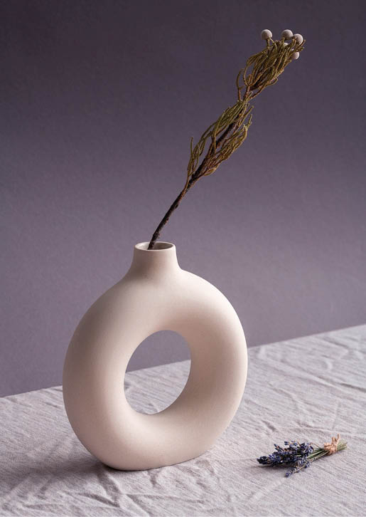 White handmade ceramic ring-shaped vase with dried wildflowers in it and small bunch of lavender lying near by on table covered with linen cloth