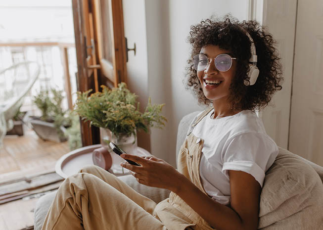 attractive dark-skinned lady looks at camera smiling broadly while sitting on sofa in light room. in her hands modern black gadget. girl in headphones, glasses and casual summer clothes.